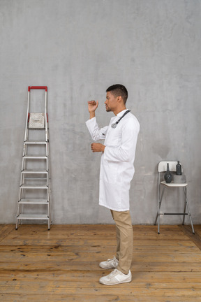 Vista lateral de un joven médico de pie en una habitación con escalera y silla mostrando el tamaño de algo