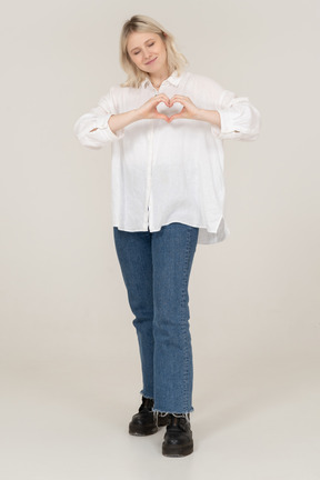 Front view of a blonde female in casual clothes showing a heart gesture with her eyes closed