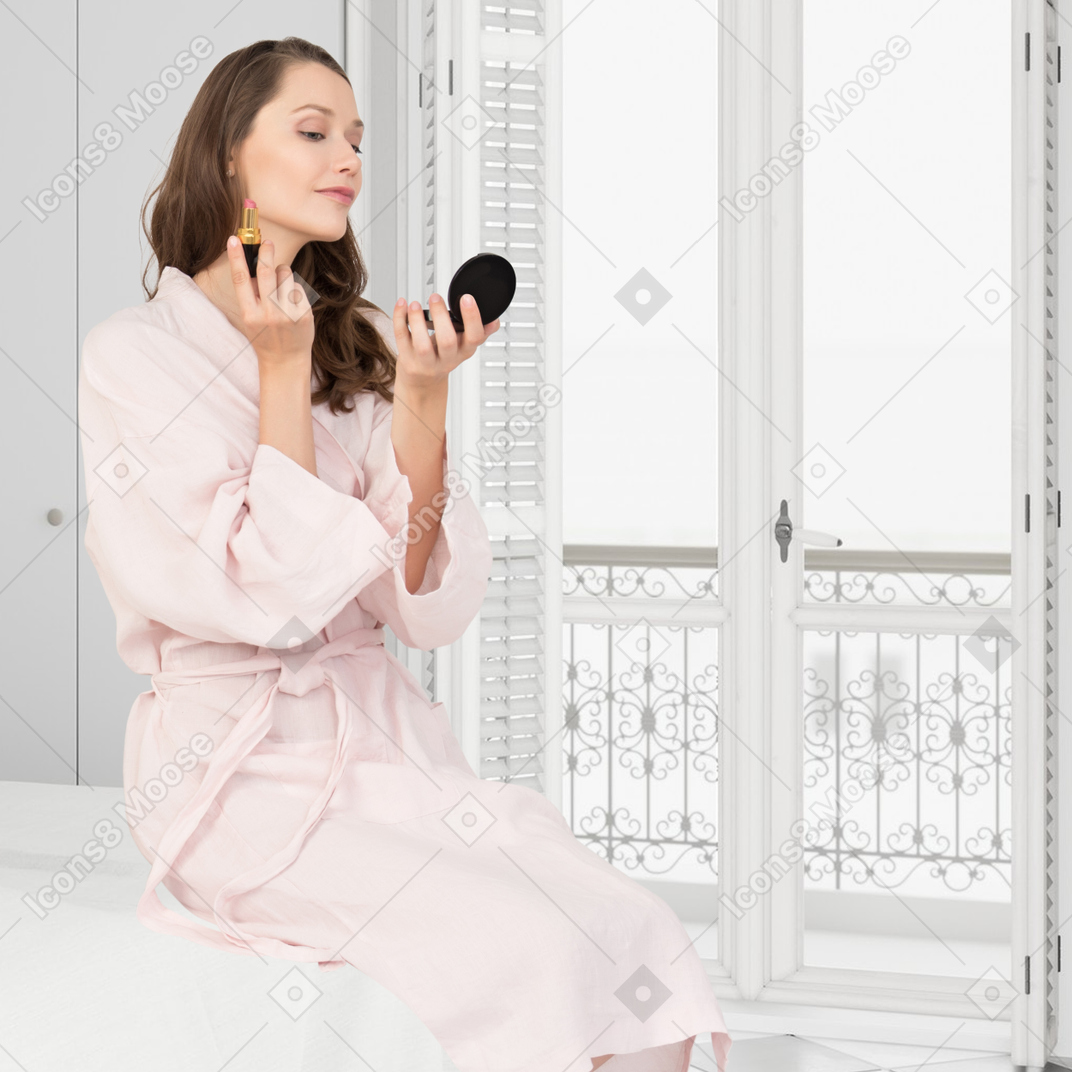 A woman in a bathrobe is holding a compact mirror and a lipstick
