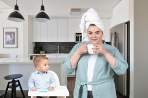 Woman with towel on her head drinking from cup while her baby sitting next to her