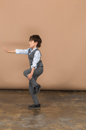 Vista lateral de um menino de terno cinza de pé em uma perna e mostrando o gesto de parada