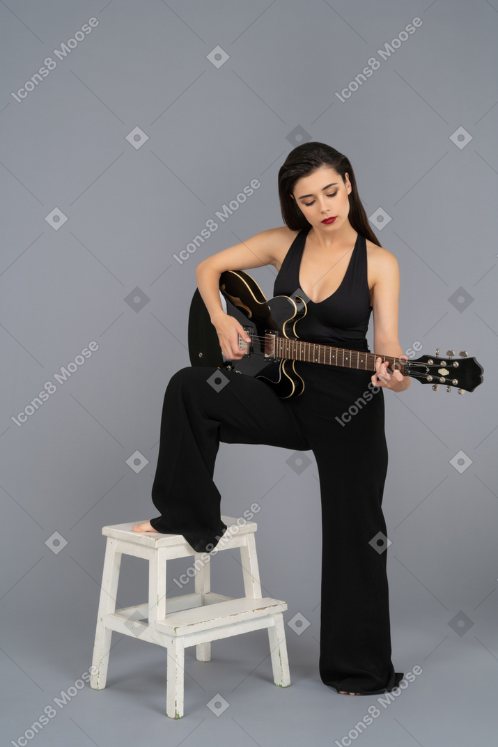 Bela jovem tocando um violão preto, mantendo a perna em um banquinho