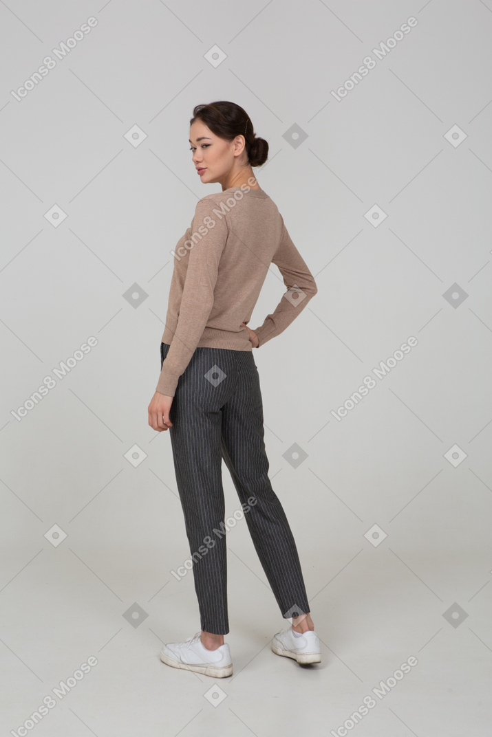 Three-quarter back view of a sly female in pullover and pants putting hand on hip
