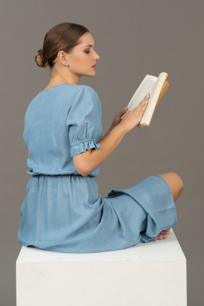 Vista laterale posteriore di una giovane donna seduta su un cubo e leggendo un libro