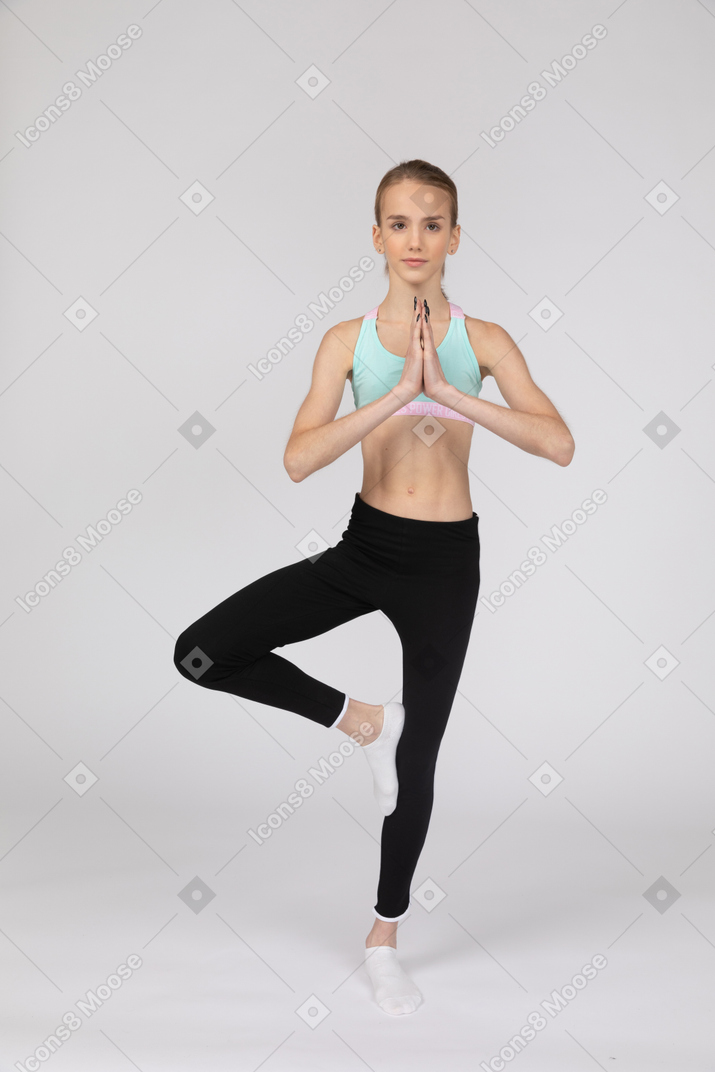 Front view of a teen girl in sportswear balancing on one leg and holding hands together