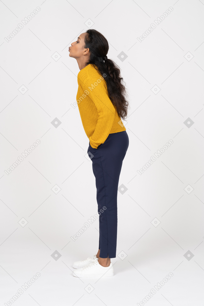 Side view of a girl in casual clothes standing with hands in pockets and blowing a kiss