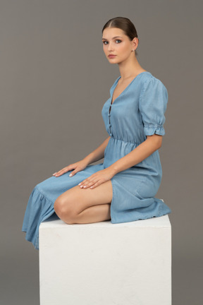 Side view of young woman in blue dress sitting on a cube