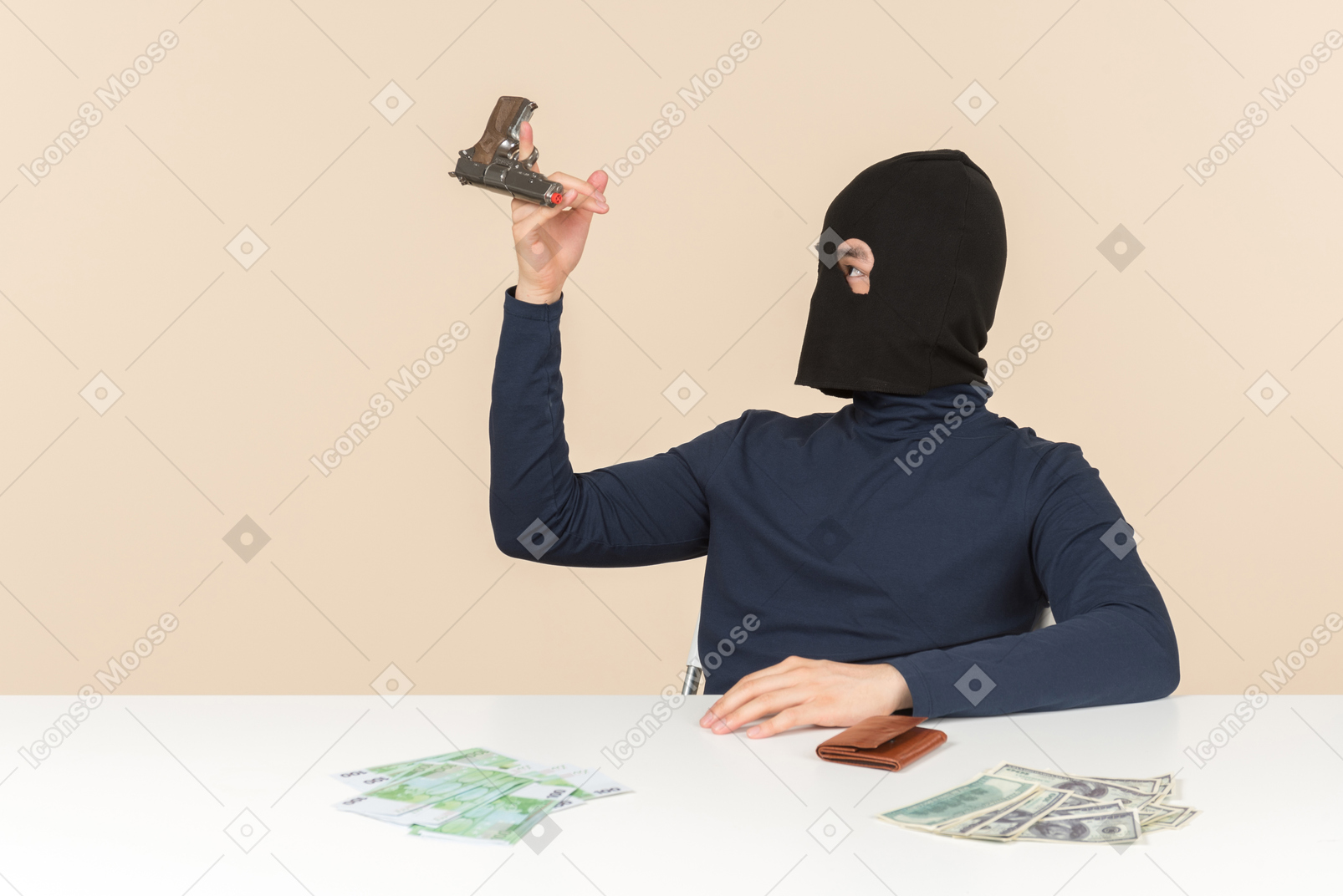 Hacker in balaclava sitting at the table with money bills on it and playing with a gun