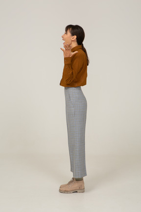 Vista lateral de uma jovem asiática surpresa de calça e blusa, levantando as mãos