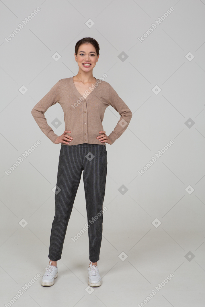 Vista frontal de una mujer sonriente en jersey y pantalones poniendo las manos en las caderas