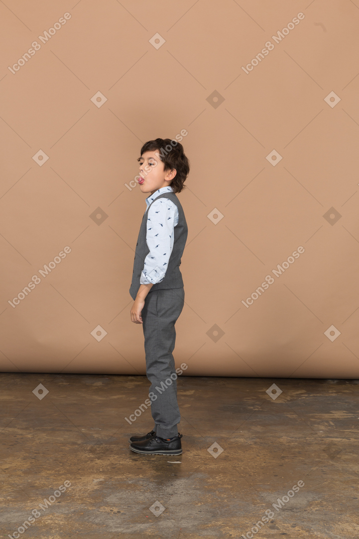 Vue latérale d'un garçon mignon en costume gris montrant la langue