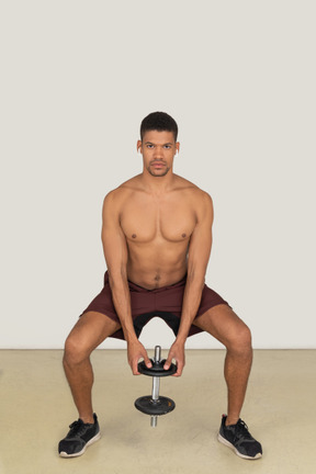 A frontal view of the muscular young guy dressed in red shorts squatting and holding the dumbbell