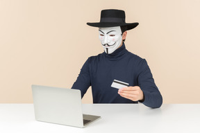 Hacker wearing vendetta mask sitting at the laptop and holding bak card
