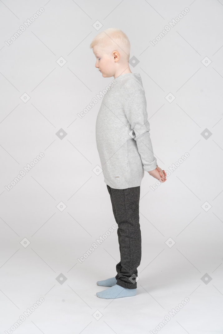 Um garotinho loiro em roupas casuais em pé e escondendo as mãos atrás