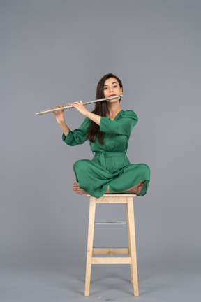 Toute la longueur d'une jeune femme jouant de la clarinette assise, les jambes croisées sur une chaise en bois