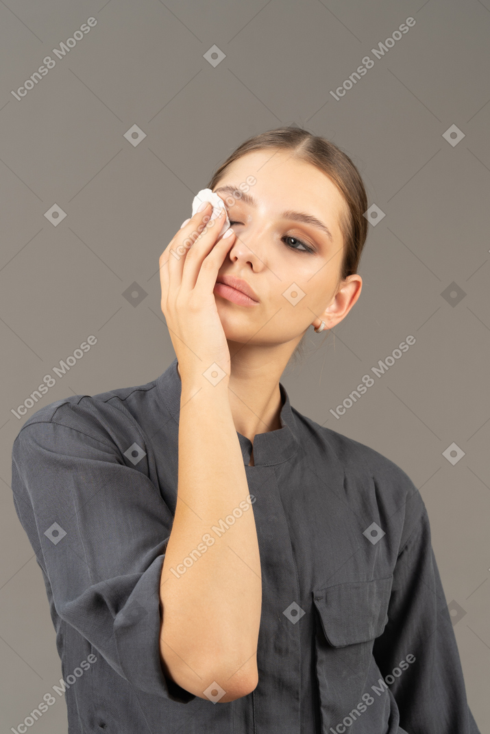 Vista frontal de uma jovem com um macacão removendo a maquiagem dos olhos