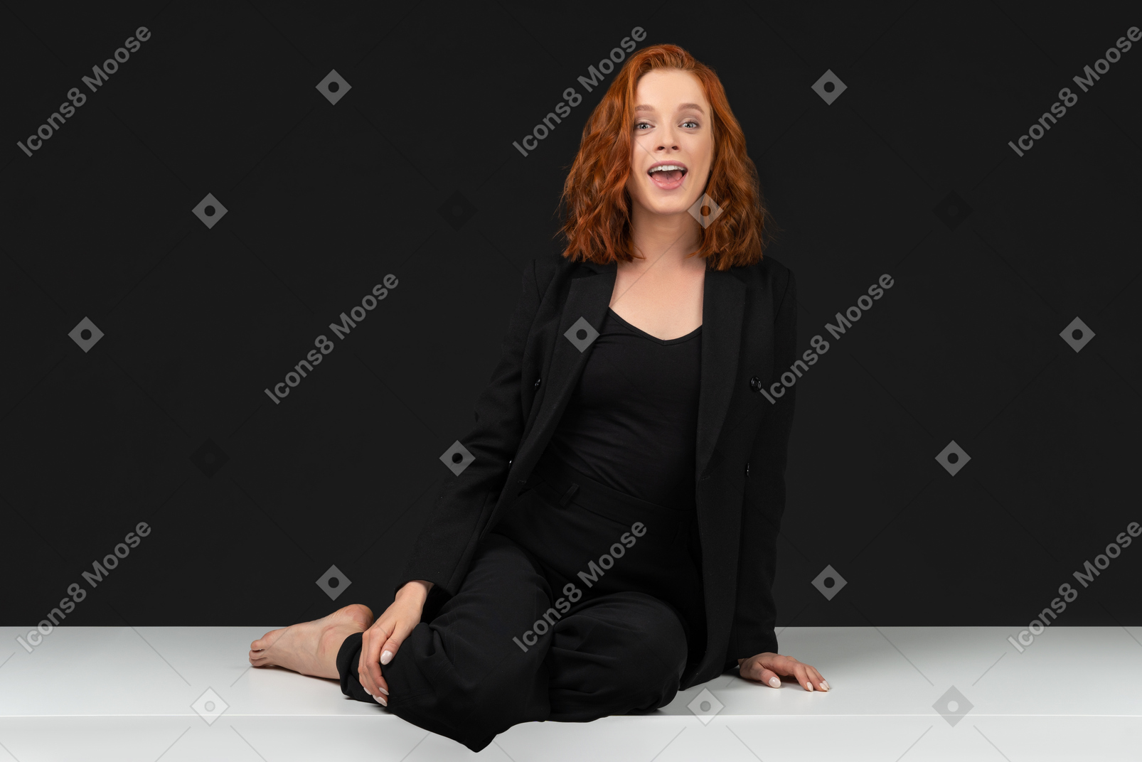 A frontal view of the cute red haired girl dressed in black and sitting on the white table
