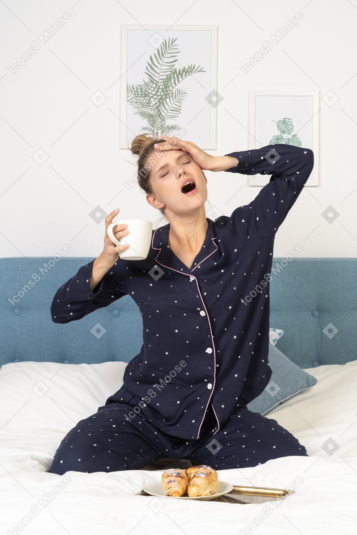 Front view of a yawning young lady in pajamas holding a cup of coffee while sitting in bed
