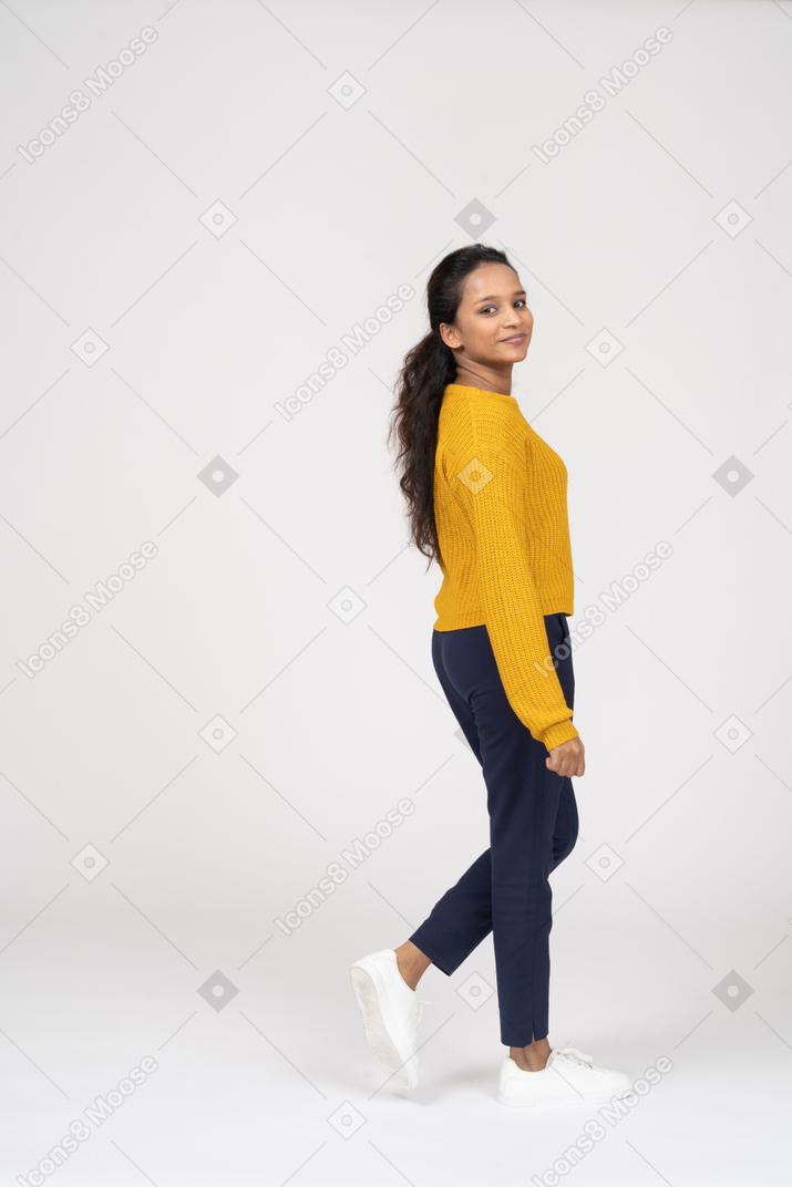 Vista lateral de uma garota feliz em roupas casuais andando e olhando para a câmera