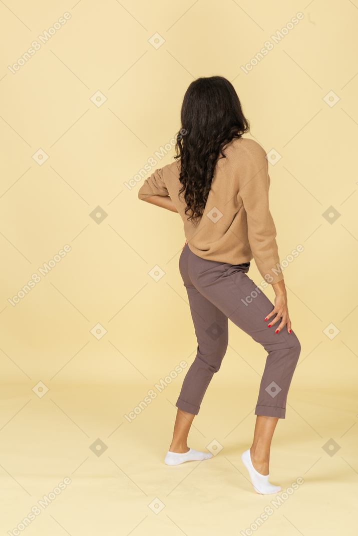 Three-quarter back view of a dark-skinned young lady putting hand on hip while looking aside