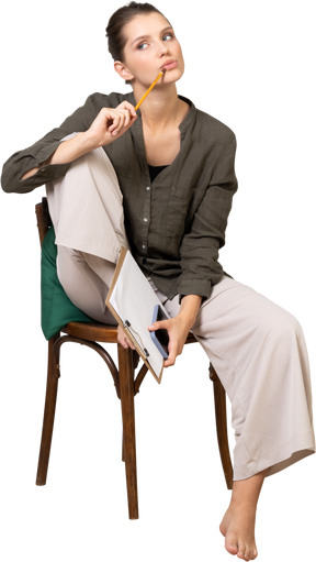 Vista frontal de uma jovem pensativa, vestindo roupas de casa, sentada em uma cadeira e fazendo anotações
