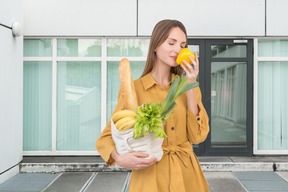 A woman in a yellow dress holding groceries bag and smelling bell pepper