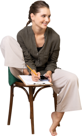 Front view of a smiling young woman wearing home clothes sitting on a chair and making notes