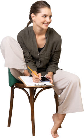 Vue de face d'une jeune femme souriante portant des vêtements de maison assise sur une chaise et prenant des notes