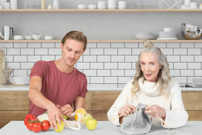 An older woman knitting and a man taking out fruits from a bag in the kitchen