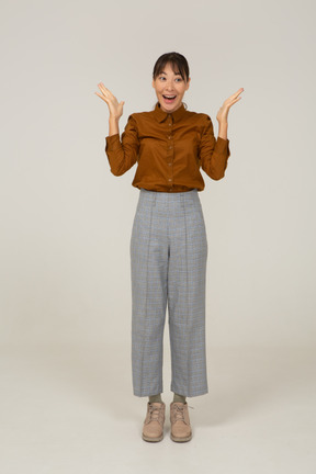 Front view of a surprised young asian female in breeches and blouse raising hands