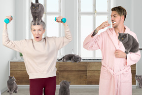 Hombre cepillándose los dientes y su madre haciendo ejercicio junto a él rodeado de gatos
