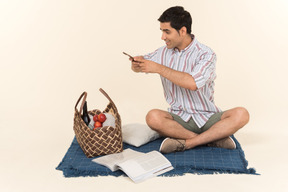 Young caucasian guy sitting on blanket and making a photo of picnic basket