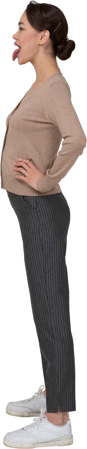 Vista lateral de una joven en jersey y pantalones poniendo las manos en las caderas y mostrando la lengua