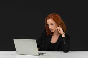 A frontal view of the cute young woman sitting at the table with laptop and touching her hair