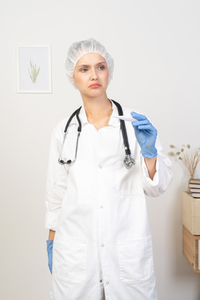 Vista frontal de una joven doctora perpleja con estetoscopio sosteniendo termómetro