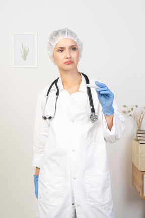 Front view of a perplexed young female doctor with stethoscope holding thermometer