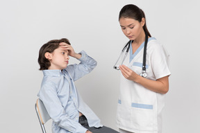 Ärztin, die auf thermometer schaut, während kinderpatient seine stirn berührt