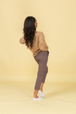 Vista posterior de tres cuartos de una mujer joven de piel oscura que pone las manos en las caderas y se inclina hacia atrás