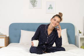 カップを持って電話をしているベッドに座っているパジャマの若い女性の前