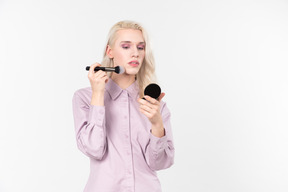 Young blond-haired person in a pastel violet shirt, doing their makeup