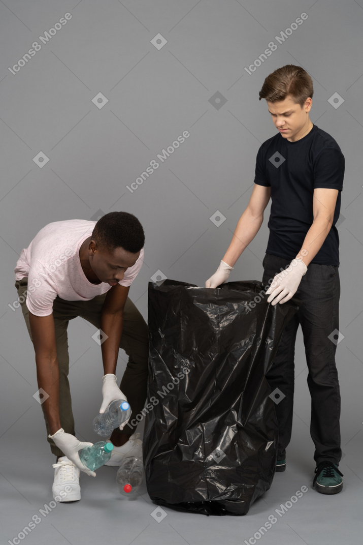 Young man helping his friend to put plastic bottles into a trash bag