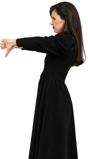 Vista lateral de uma jovem insatisfeita de vestido preto com os polegares para baixo