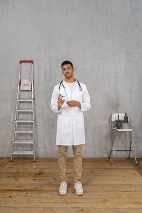 Front view of a young doctor standing in a room with ladder and chair showing a size of something