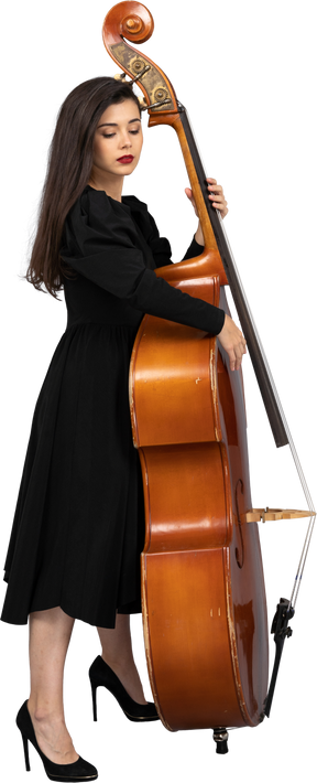 Vista lateral de un joven músico serio en vestido negro tocando su contrabajo