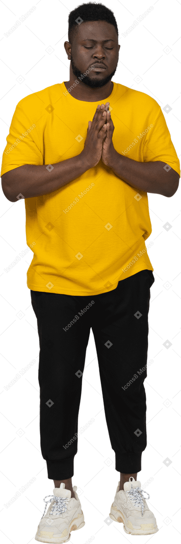 Front view of a praying young dark-skinned man in yellow t-shirt holding hands together