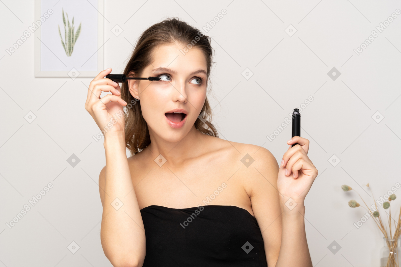 Front view of a surprised young woman wearing black top applying mascara