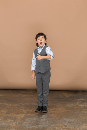 Вид спереди на симпатичного мальчика в сером костюме, показывающего большой палец вверх