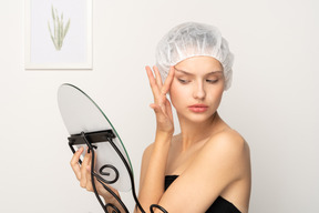 Young woman tightening her face skin while looking in the mirror