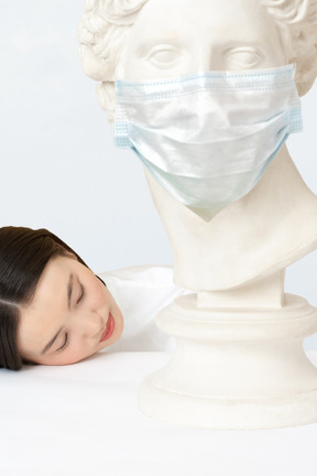 A woman laying her head on a table next to a bust of a woman in face mask