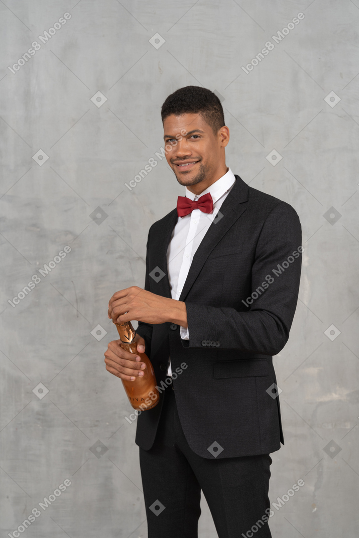 Man in suit and bow tie standing and opening a bottle of champagne